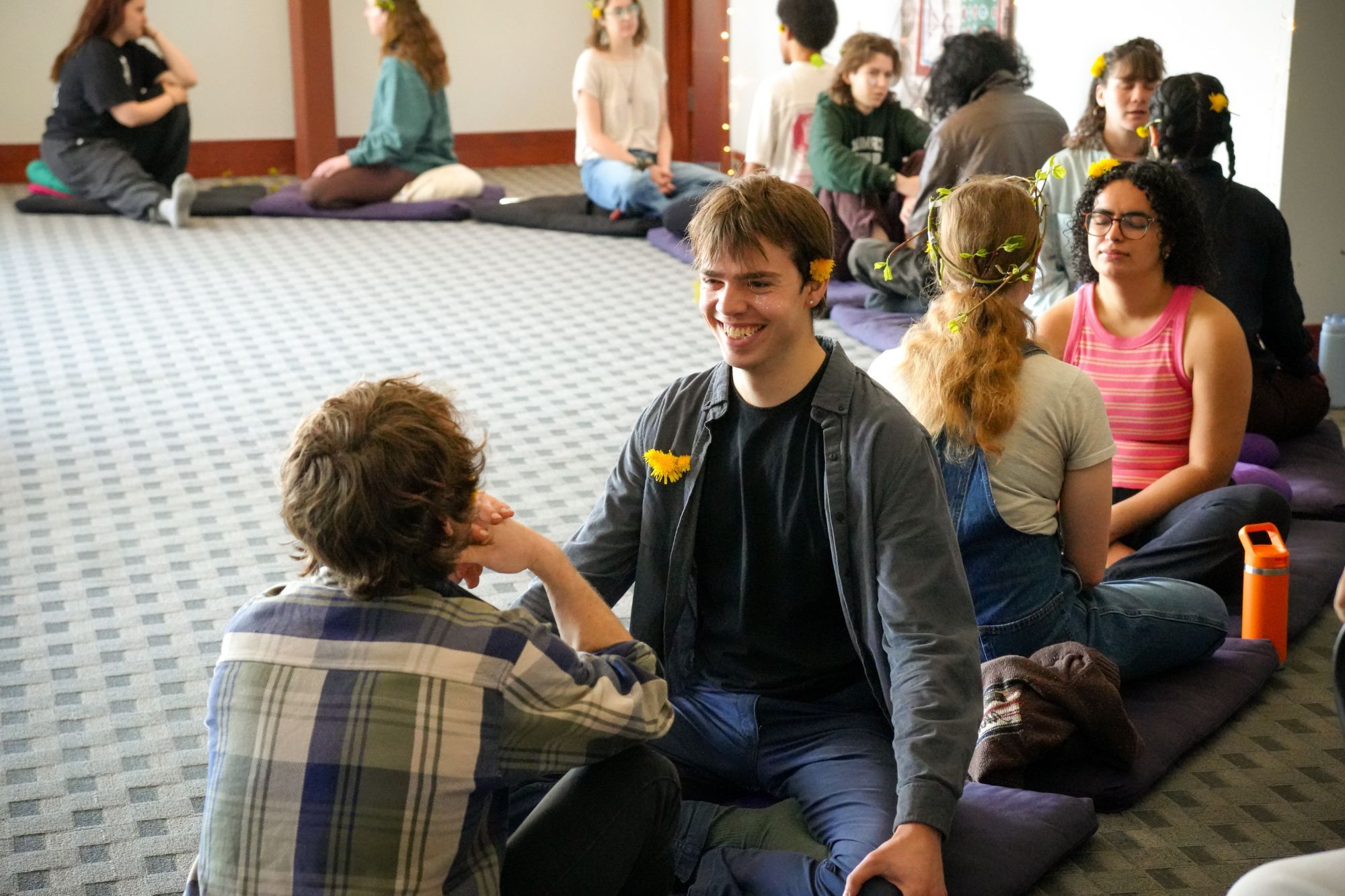 Young people sit on cushions in pairs, talking and listening. In front, a smiling young man with a dandelion behind his ear faces a friend, and flower crowns and dandelions decorate the people around them.