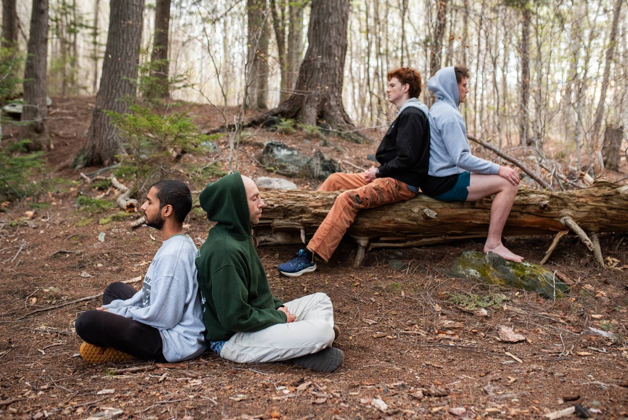 Two pairs of people sit back to back in the forest, eyes closed or gazing ahead, some on the ground and some on a fallen log.
