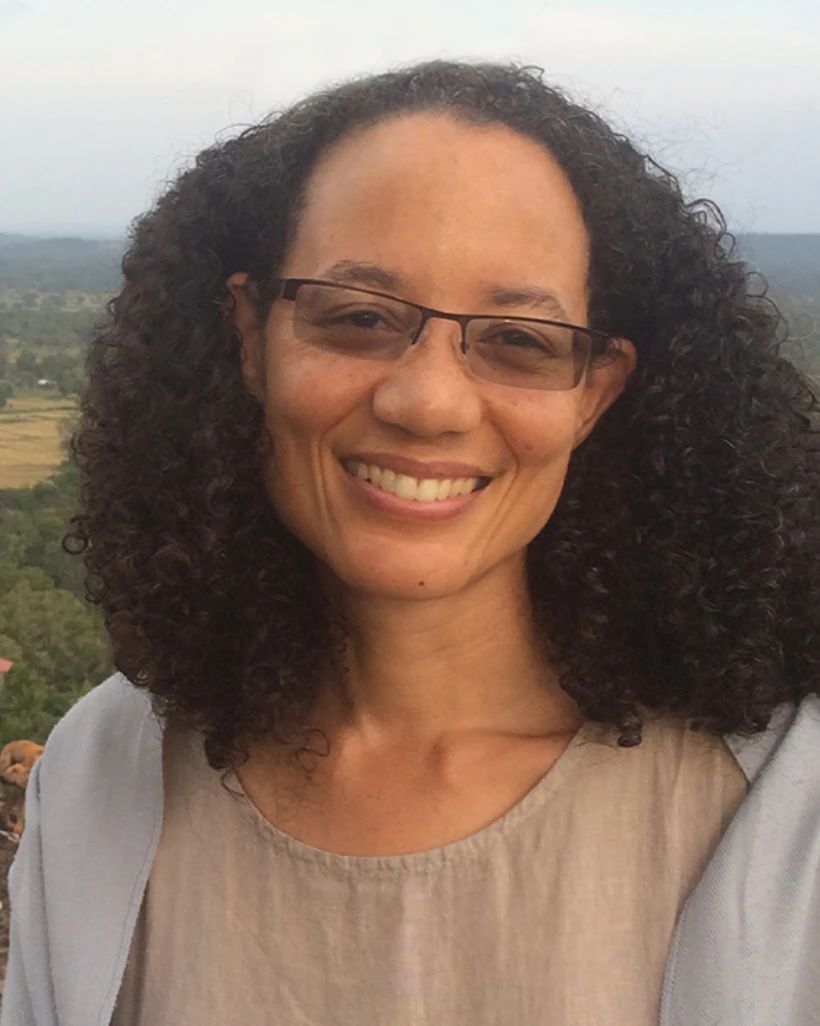 Kaira Jewel Lingo, smiling, with curly hair and glasses, outdoors with a wide landscape behind her.