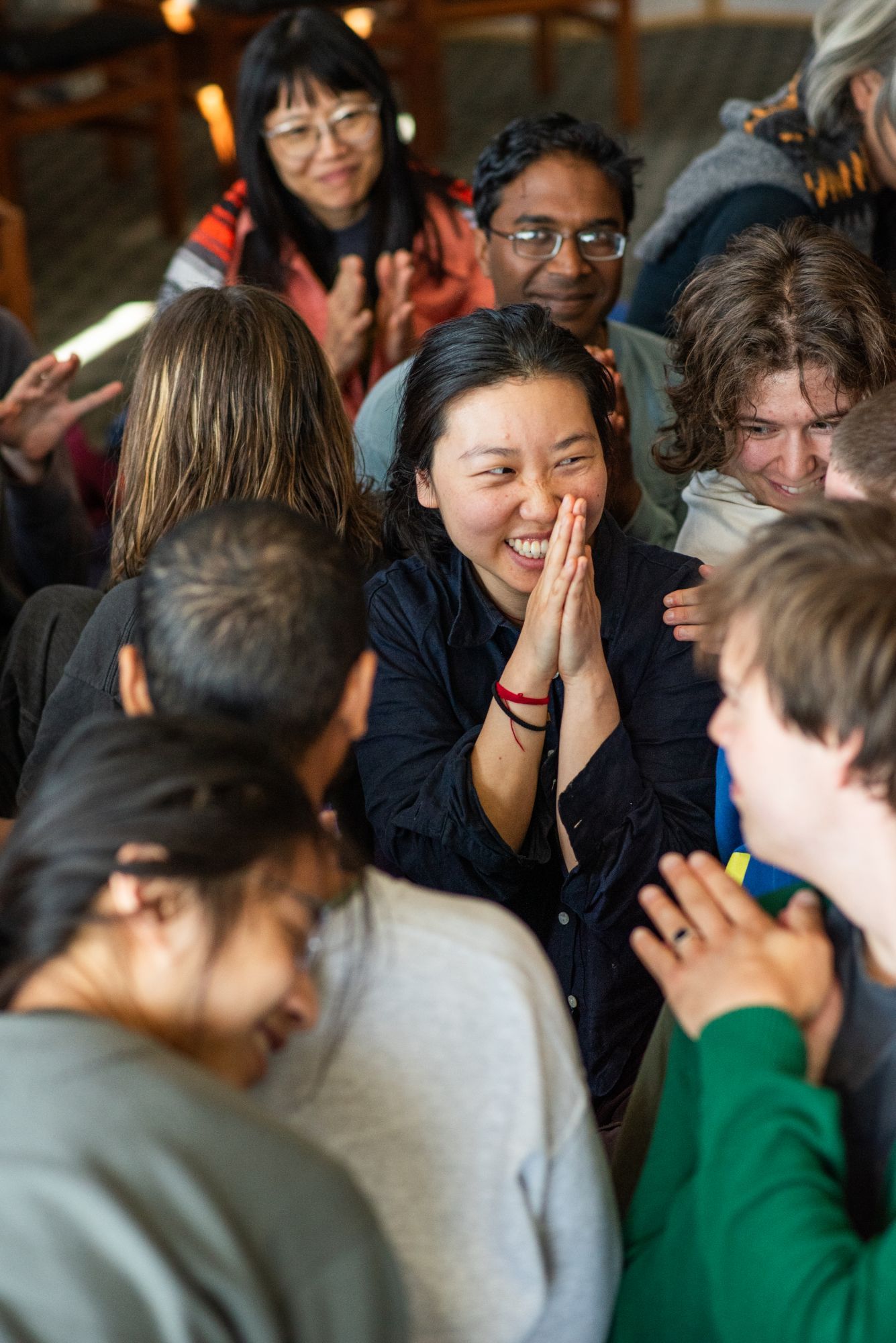 A smiling person with palms pressed together, surrounded by laughing friends.
